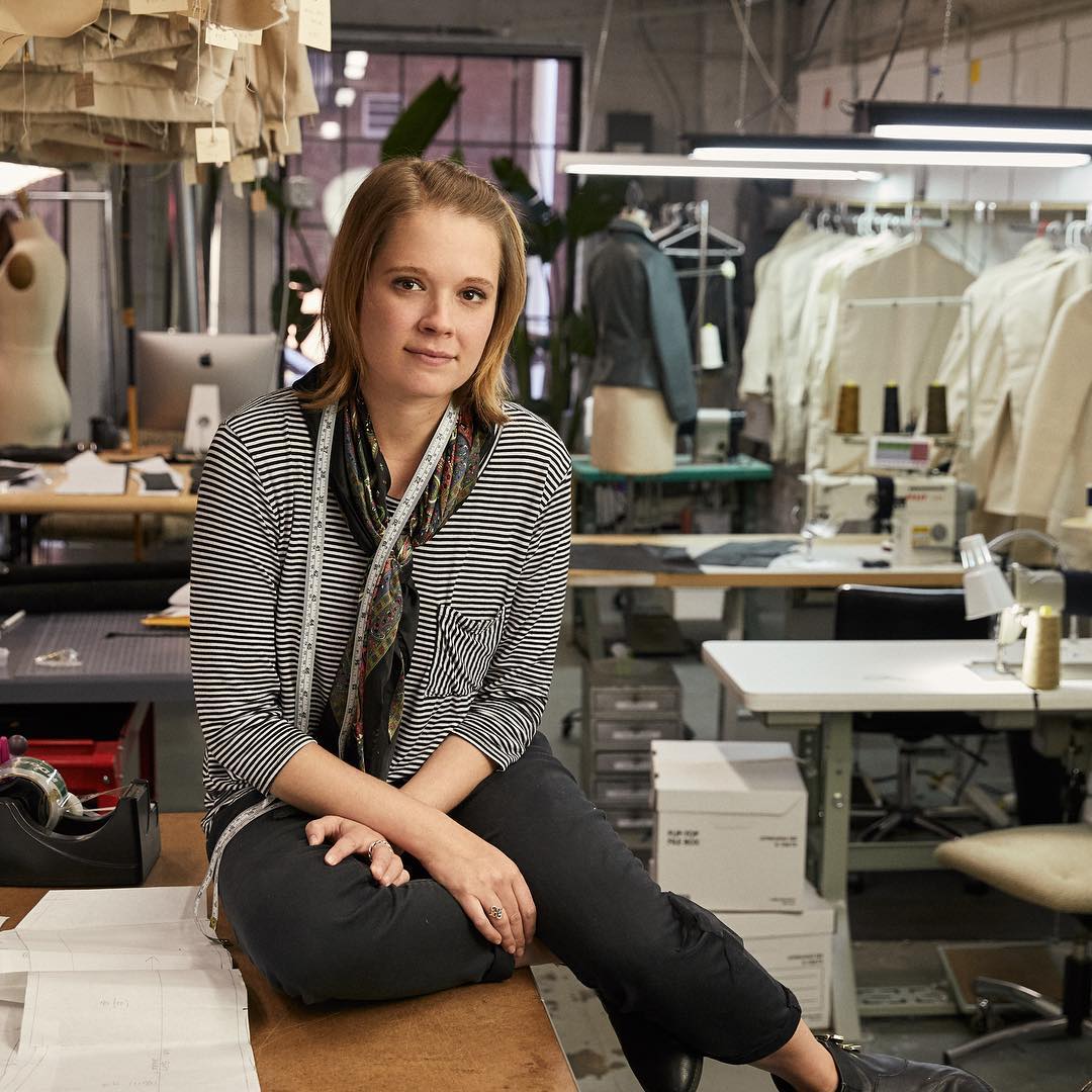 Annie Masarie sitting on table with paper patterns around her, garments and suit jackets hanging in the background, Clothing Measuring Tape around her neck, mannequins and sewing machines in the background for sizing and fitting