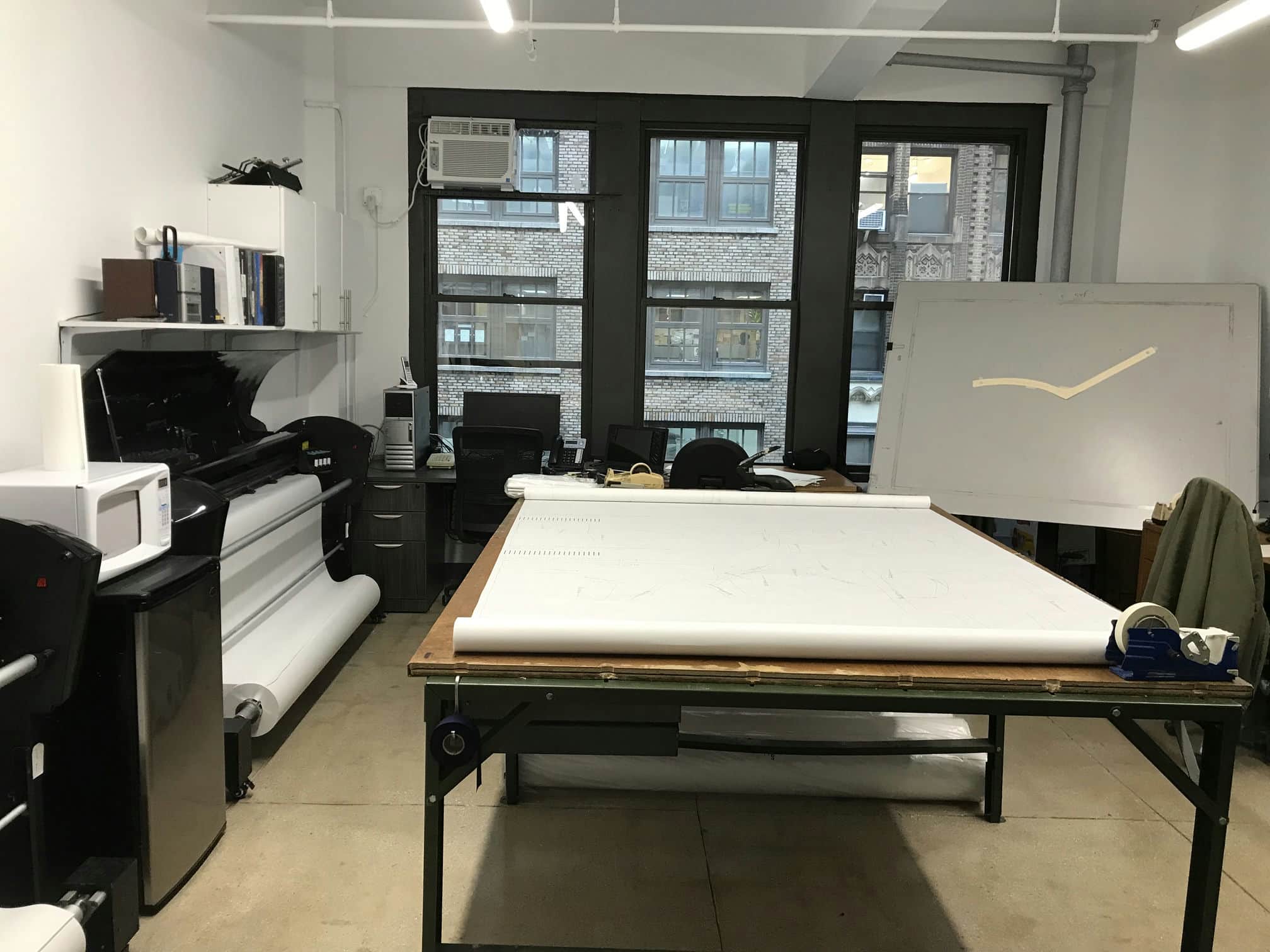 Wide shot of the Top Notch Pattern office showing plotters and printers, a marker on the table in the middle ready for garment cutting and samples, a paper pattern on Lectra digitizer table, background of skyscraper in garment district of NYC